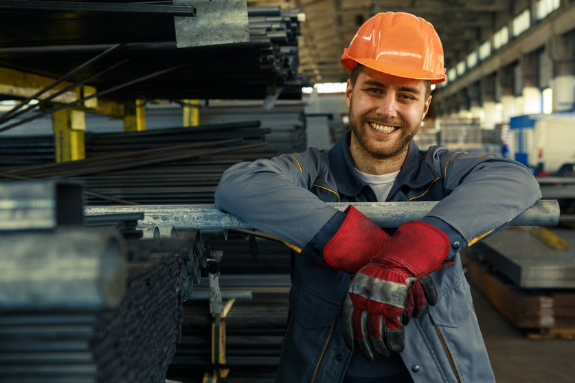 Cheerful young worker in hardhat and uniform smiling to the camera while working at the storage copyspace metal steel metalworking factory production concept