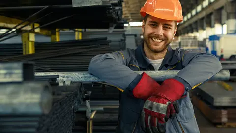 Cheerful young worker in hardhat and uniform smiling to the camera while working at the storage copyspace metal steel metalworking factory production concept